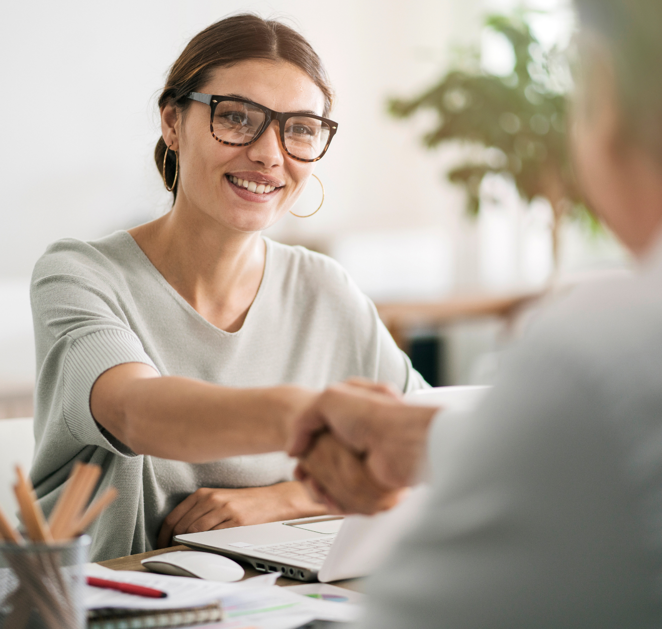 A woman wearing glasses is smiling and shaking hands over their desk.