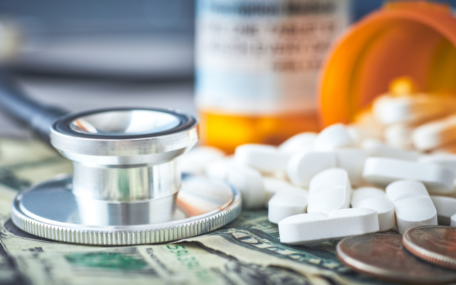 A stethoscope is beside a prescription bottle with medicine poured out over the top of money.