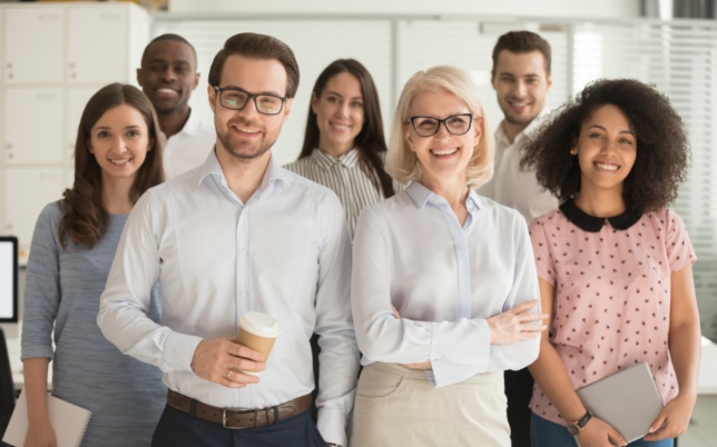 Seven employees are standing in a group smiling for a photo at work.