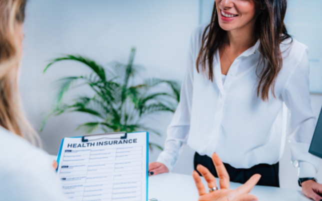 A woman is holding a paper that says health insurance and talking to another woman who is smiling.