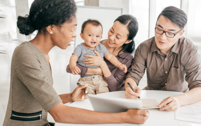 A woman is talking to a couple. The lady and man are sitting together, the lady holding their baby and the man is signing papers.