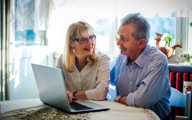 An elderly man and woman are sitting behind a laptop computer, smiling and talking.