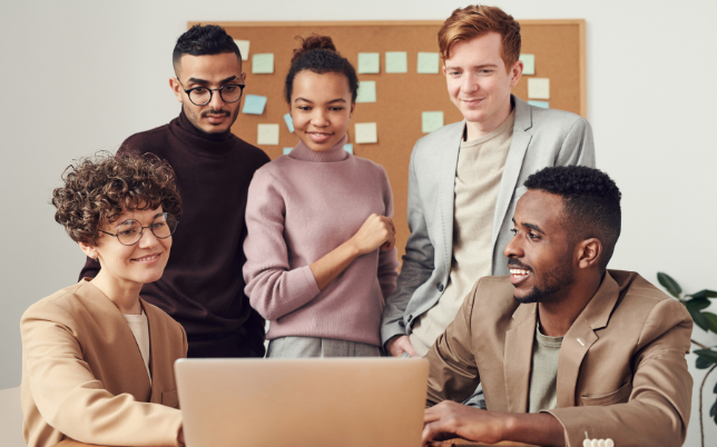 A small group of five employees is surrounded by a laptop talking and smiling.
